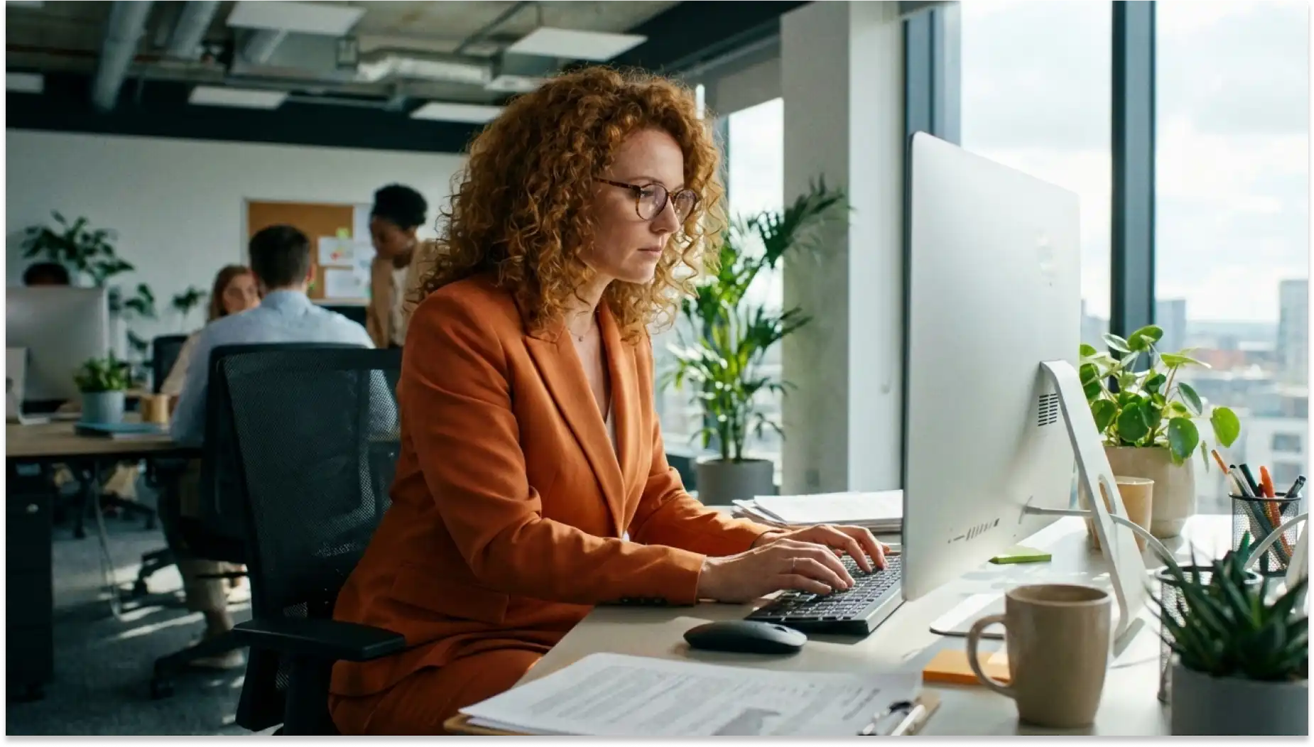 Mulher de cabelos cacheados e vestindo blazer laranja, representando mulheres no mercado de trabalho. trabalha em escritção moderna com computador e plantas ao fundo.
