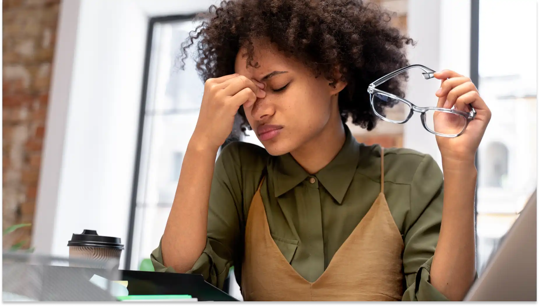 Mulher jovem com expressão de dor de cabeça, segurando os óculos e com a mão na testa, em ambiente de escritório ou estudo, representando afastamento no trabalho.