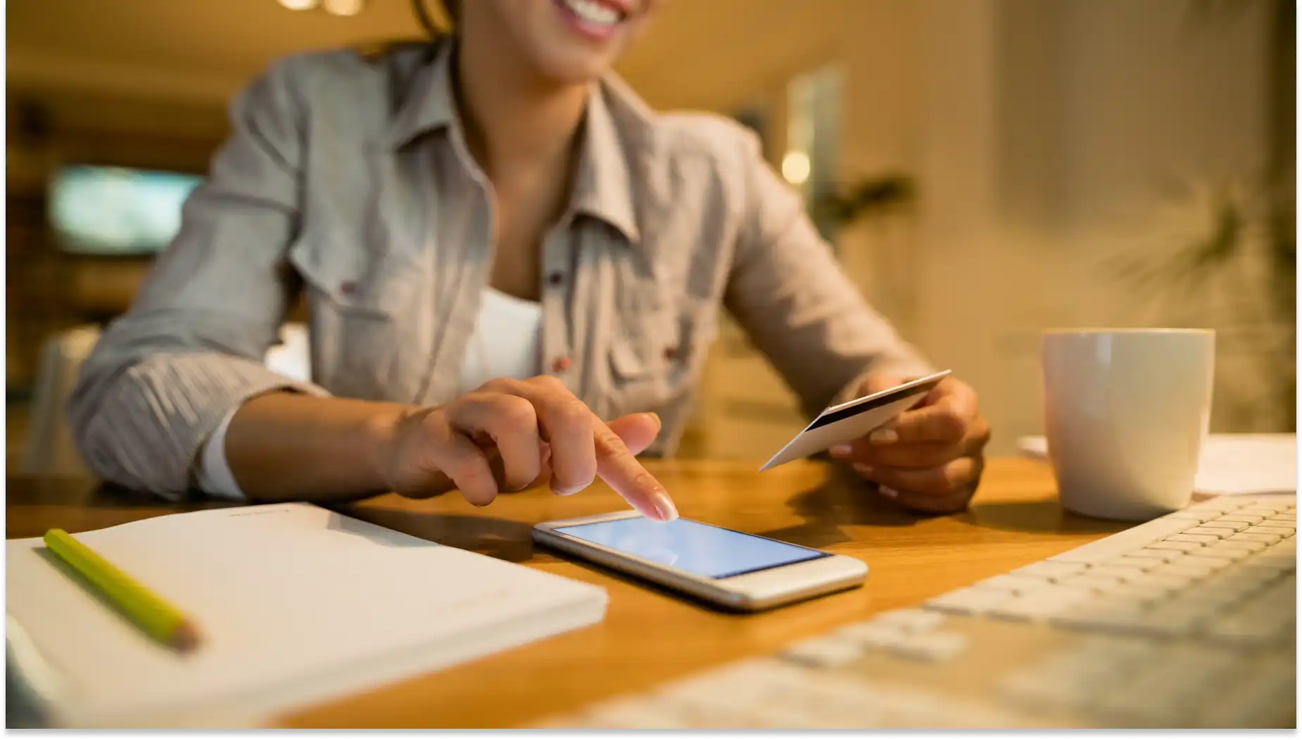 Mulher usando smartphone e cartão de crédito em ambiente aconchegante, com caderno e café na mesa, realizando compras online com segurança e comodidade para representar inclusão financeira.