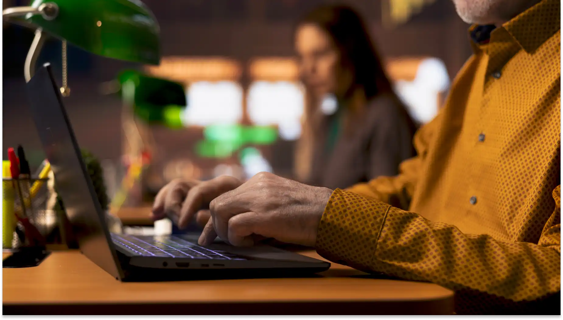 Pessoa usando notebook em ambiente de escritório moderno com outras pessoas ao fundo, ambiente de força de trabalho colaborativo e tecnológico.