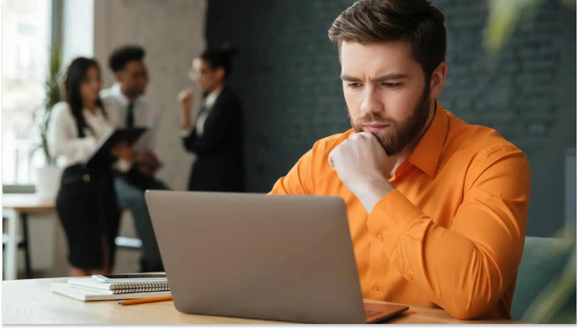 Homem pensativo usando laptop em escritório, enquanto colegas ao fundo conversam, representando trabalho, foco em liberar informes de rendimentos de 2025.