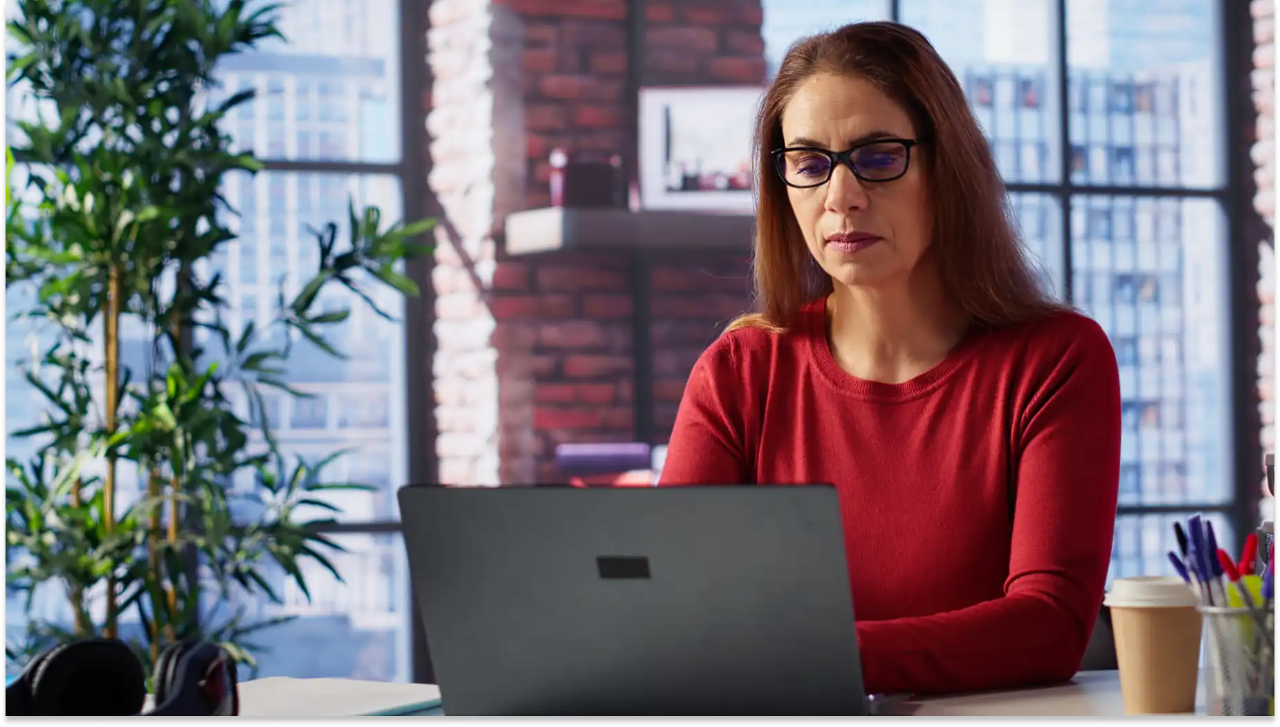 Mulher de óculos trabalhando no computador em um escritório moderno, representando trabalho em feriados, com decoração contemporânea e plantas ao fundo, refletindo concentração.