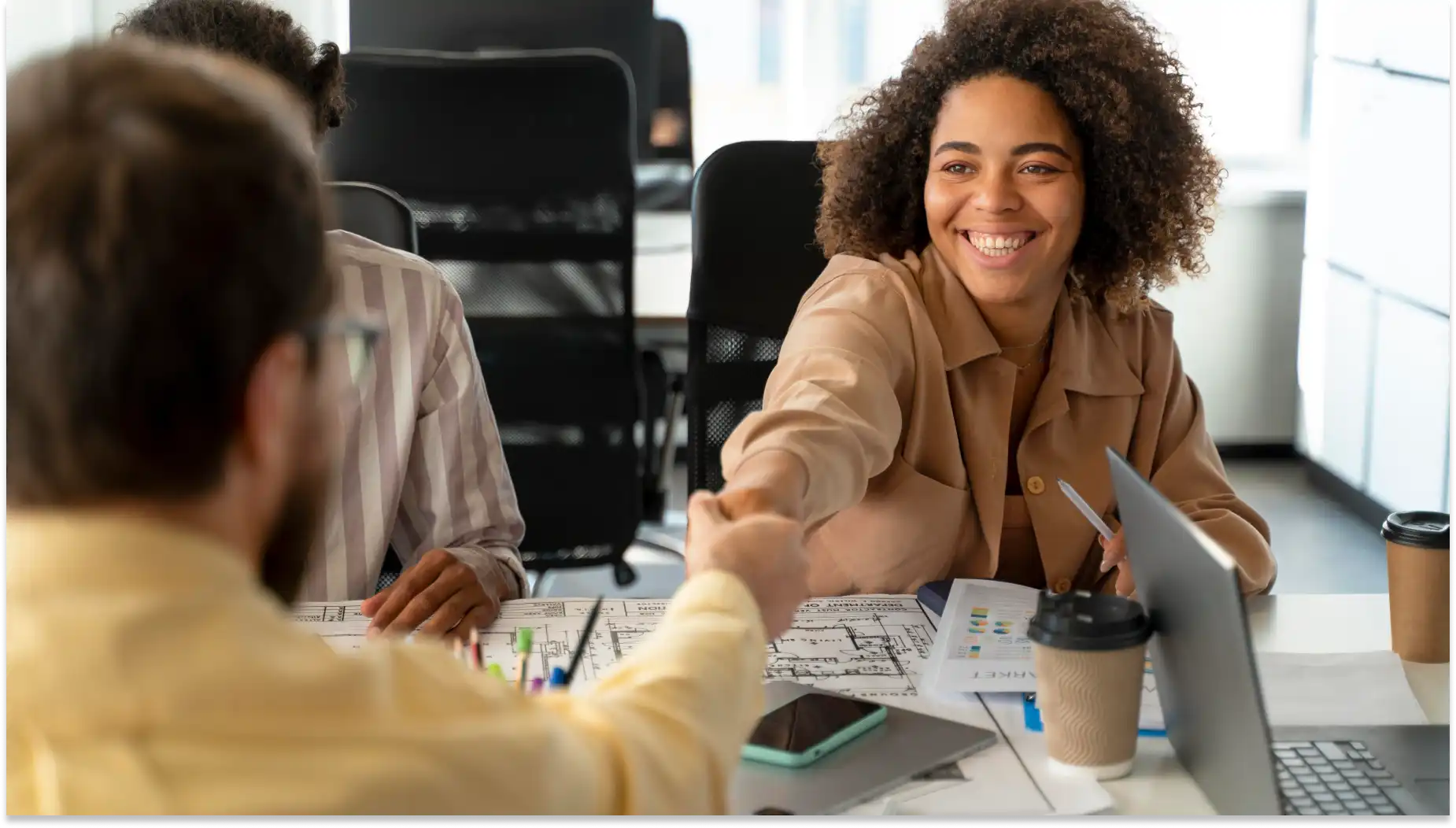 Profissional feminina sorridente contratando profissional simbolizando empregos formais em reunião no escritório, com materiais de trabalho, laptop e café na mesa.