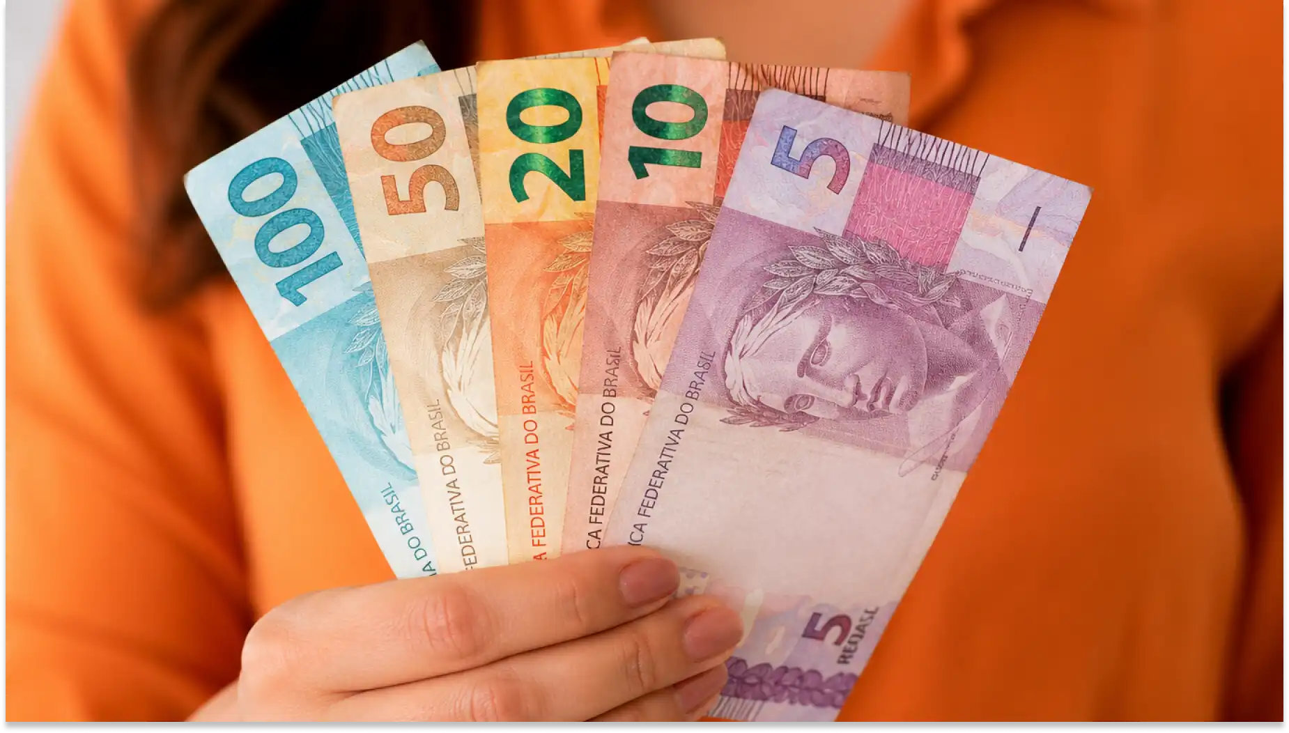 Imagem de mãos segurando diferentes cédulas de dinheiro, representando impacto do reajuste salarial na margem para CLT em 2026.