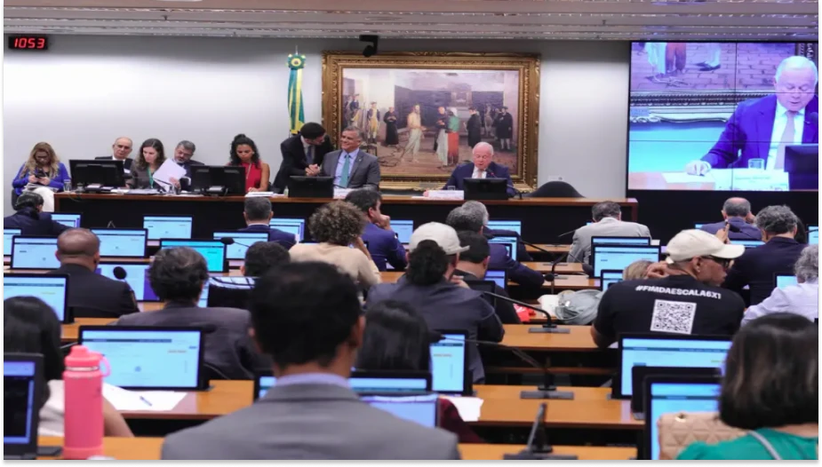 Reunião formal de profissionais e autoridades em uma sala de conferências, com apresentação de slides, computadores e uma grande tela de projeção. Ambiente de trabalho colaborativo e oficial.