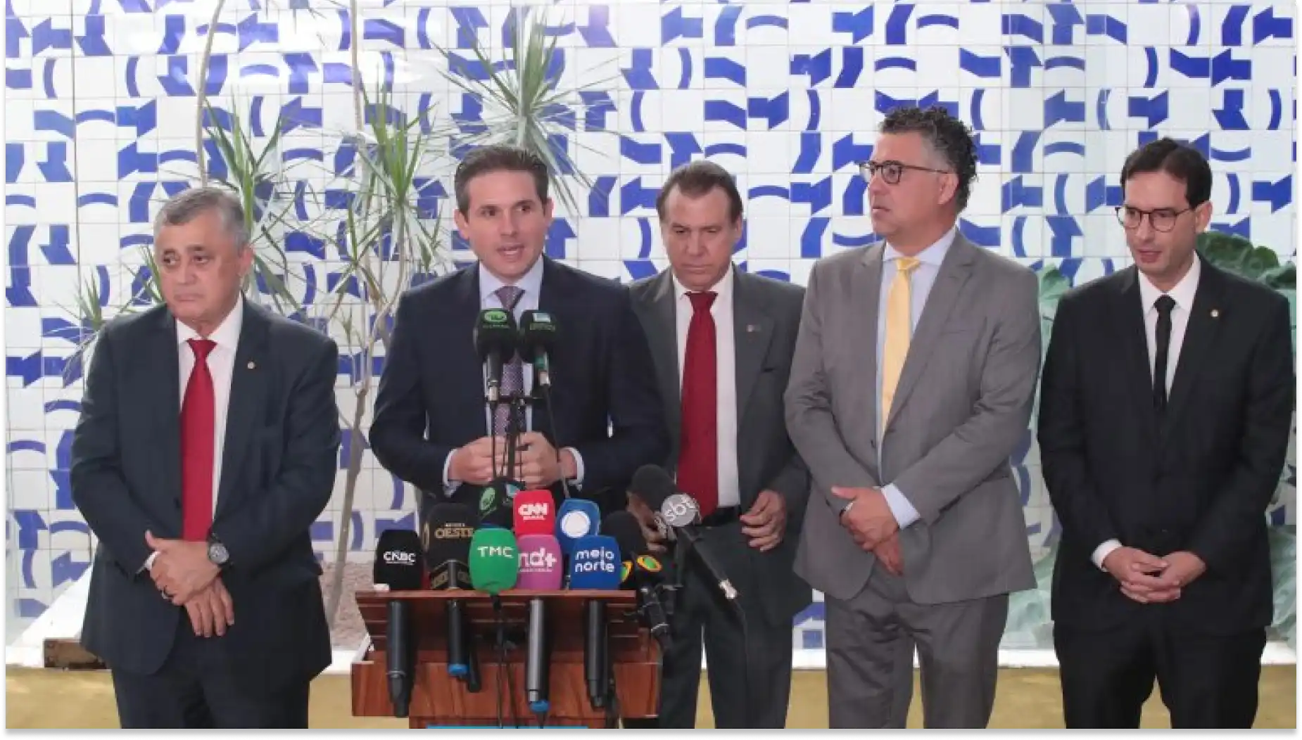 Encontro de autoridades em entrevista coletiva: cinco homens em trajes formais ficam ao lado de mesa com microfones e bandejas, diante de painel com padrão azul e branco, em ambiente externo, disutindo escala 6x1.