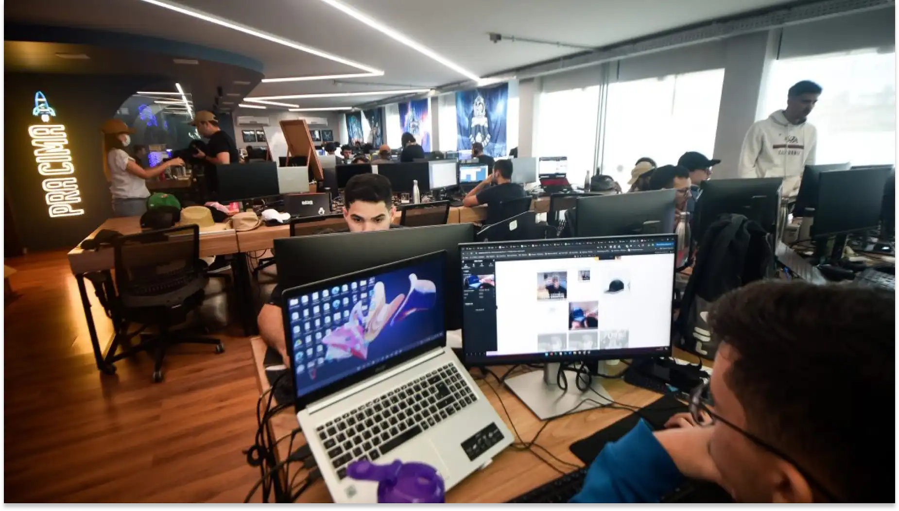Ambiente de um escritório de tecnologia com várias pessoas trabalhando em computadores, incluindo um espaço de coworking com decoração moderna. QuarkRH