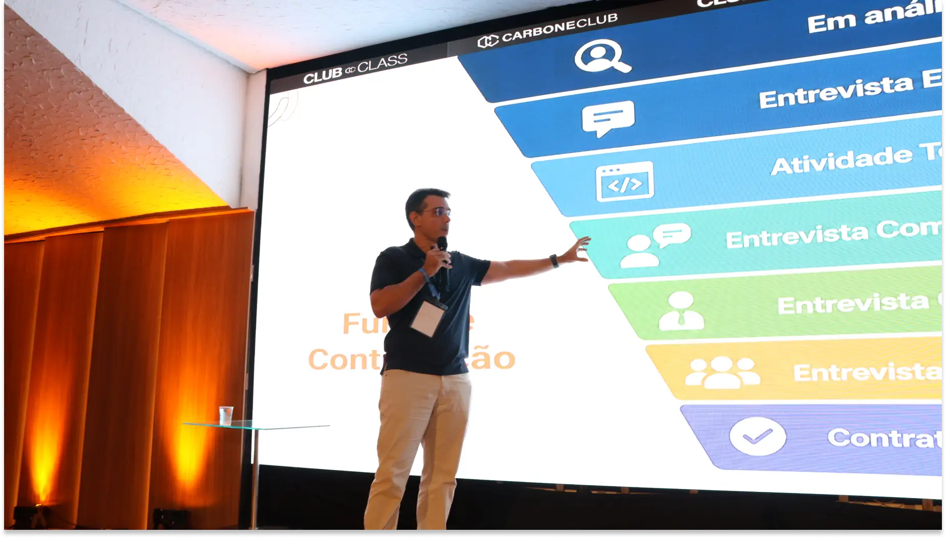 Palestrante em um palco, fazendo apresentação com microfone diante de um telão que exibe informações e ícones sobre Insights palestra “Contratação e Retenção - Carbone Class”.