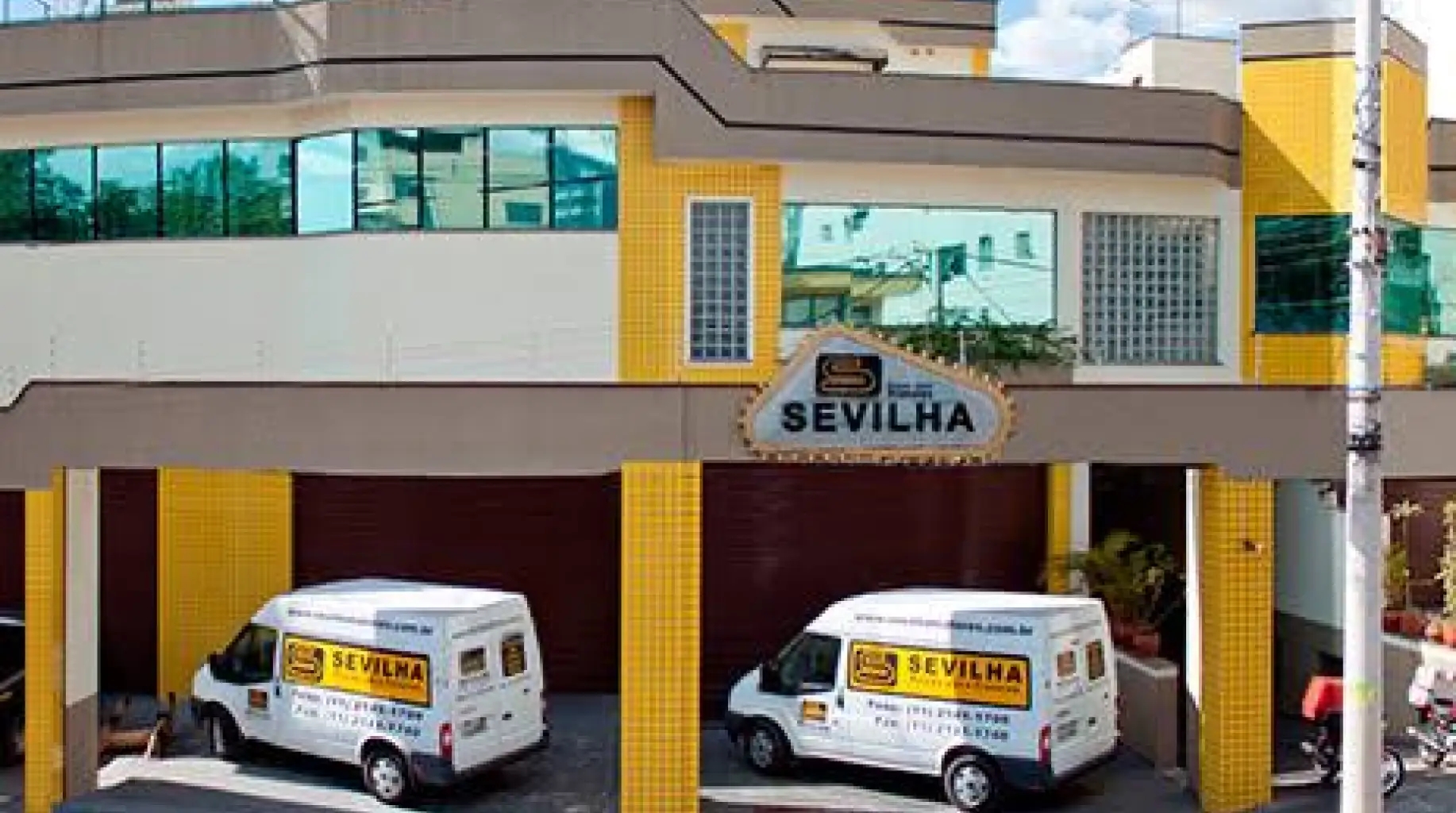 Entrada de um prédio com fachada amarela e placa “SEVILHA” acima do acesso, com duas vans brancas estacionadas na garagem, cada uma com o mesmo letreiro Sevilha.