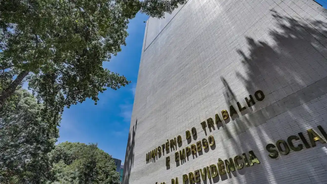 NR-1: Fachada de um prédio com o texto “MINISTÉRIO DO TRABALHO E EMPREGO” e “COLETA PREVENTIVA”, com árvores ao lado e céu azul ao fundo.