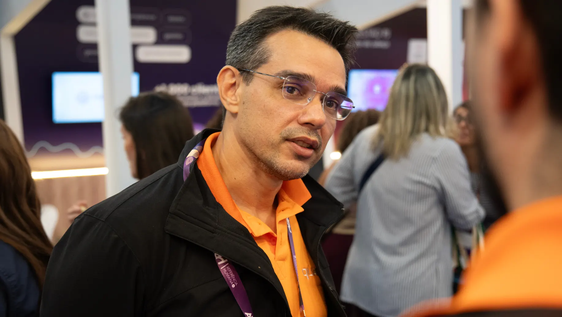 CEO da QUARKRH Gleydson Lima de evento usando crachá em um estande, em ambiente de feira corporativa, com fundo de painéis e pessoas circulando durante a CONARH 2026.