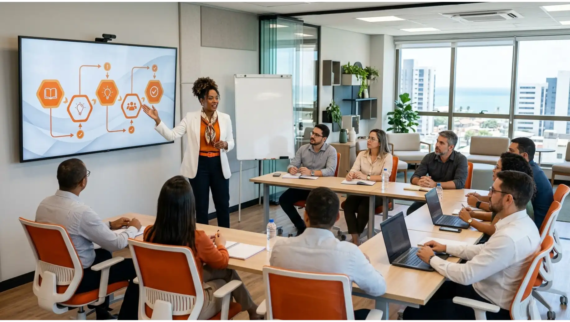 Microlearning em sala de treinamento corporativo: instrutora aponta para um quadro com fluxograma e ícones, enquanto equipes assistem sentadas ao redor de uma mesa com laptops e anotações.